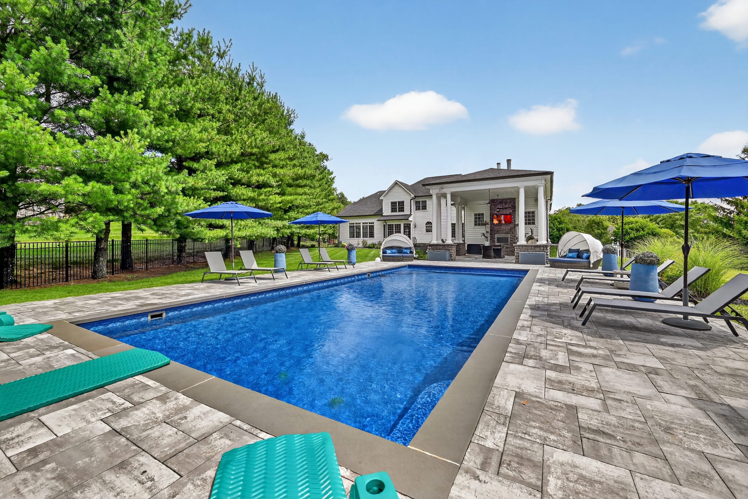 A luxurious backyard with a swimming pool, surrounded by green trees, lounge chairs, and blue umbrellas. An elegant house is visible in the background.