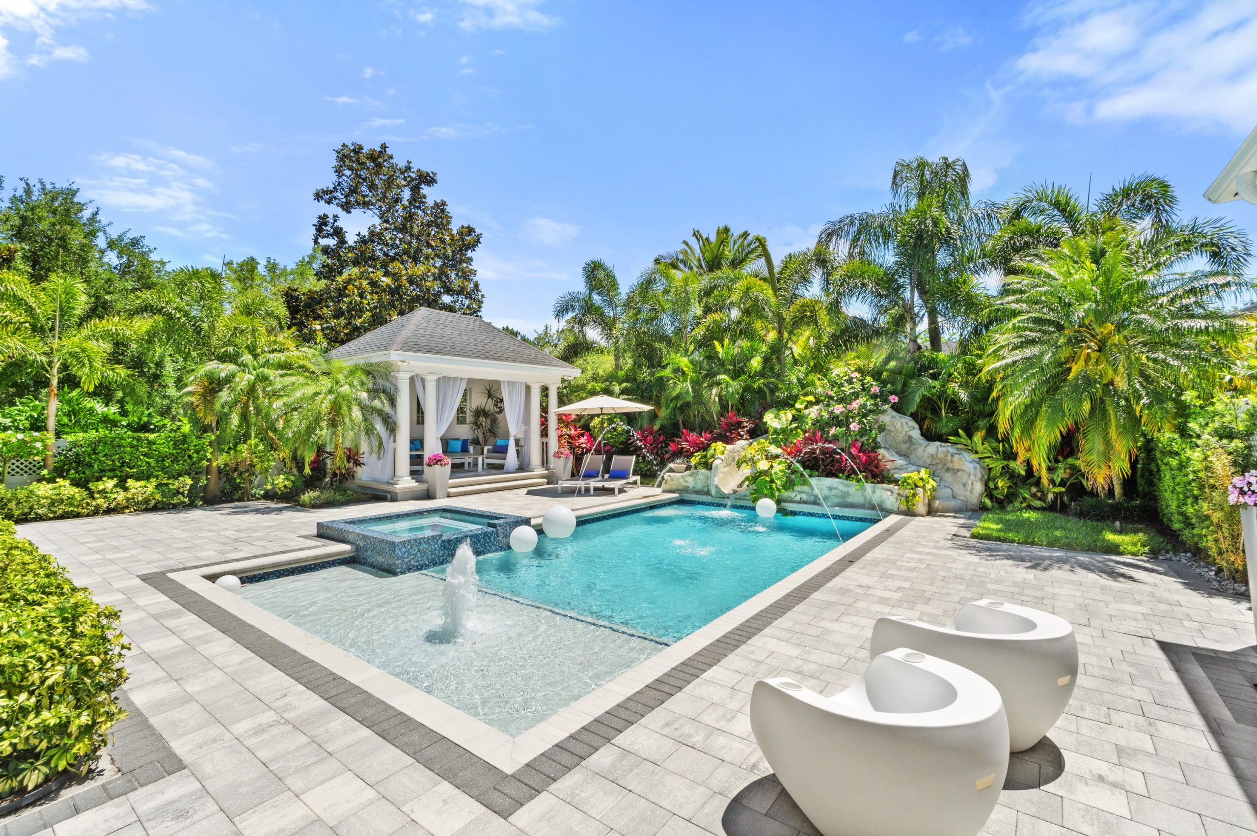A beautifully landscaped backyard featuring a swimming pool with a fountain, surrounded by tropical plants and flowers, with a gazebo and outdoor seating area.