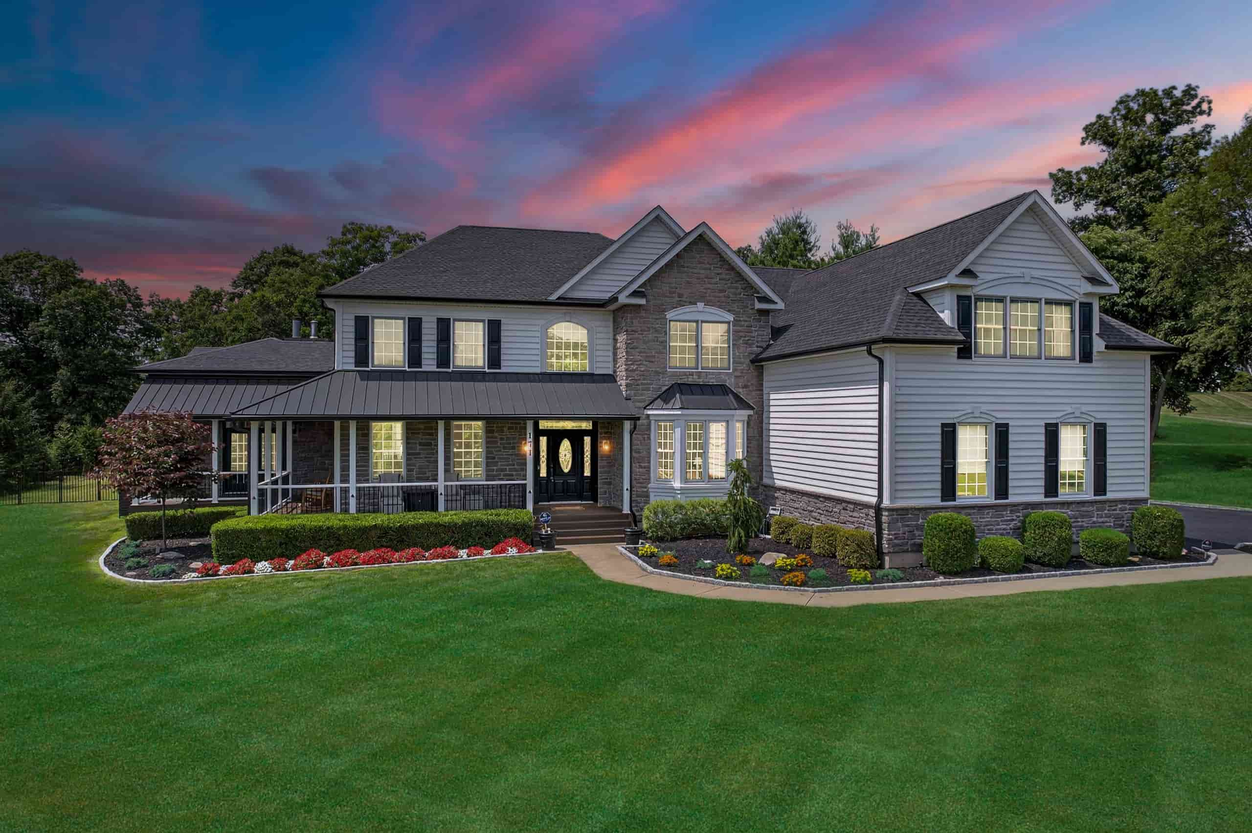 A large two-story home with a mix of stone and siding exterior, surrounded by a well-manicured lawn and colorful flower beds, under a vibrant sunset sky.